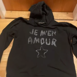 Svart hoodie med glittertext Gina Tricot - Svart hoodie från Gina Tricot med glittrig text 'JE M'EN AMOUR' och en stjärna framtill. Tröjan har huva och långa ärmar, perfekt för en chill och trendig look. Mjuk och skön, passar till många outfits.