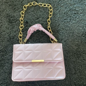 Quiltad ljusrosa axelväska med kedja - Säljer en ljusrosa quiltad axelväska med guldig kedja och handtag i matchande rosa material. Väskan har en stilren rektangulär form och stängs med ett guldfärgat spänne. Perfekt för dig som vill ha en söt och trendig accessoar till din outfit.