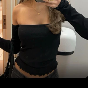Svart offshoulder topp med vågig kant - Trendig svart offshoulder topp med långa ärmar och croppad passform. Toppen har en snygg vågig kant nertill som ger en cool detalj. Perfekt för dig som vill ha en stilren men ändå edgy look.