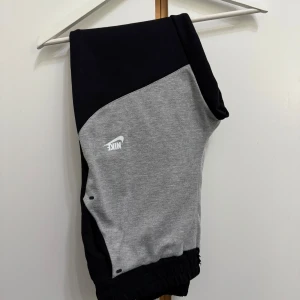  mjukisbyxor svart och grå -  mjukisbyxor i svart och grått med vit logga på låret. Byxorna har elastisk midja, muddar vid benslut och två fickor fram. Tillverkade i mjukt bomullsmaterial för maximal komfort och en sportig look.