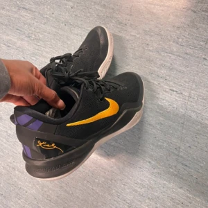 Kobe 2 basketskor  - Snygga svarta kobe 2 basketskor med mesh-ovandel, gul swoosh på sidan och lila detaljer vid hälen. Skorna har signaturdetalj i guld på hälen och svart snörning. 