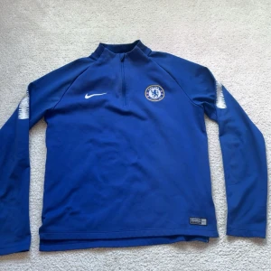 Chelsea träningströja - Chelsea FC träningströja från Nike i blått med klubbmärke på bröstet och vit Nike-logga. Långärmad modell med half zip och Dri-FIT-material som andas. Perfekt för fotbollsträning eller chill dagar. Snygga vita detaljer på ärmarna.