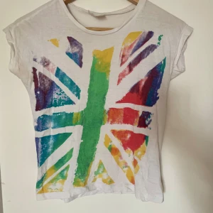 Vit t-shirt med färgglatt tryck  - Vit t-shirt med korta ärmar och en stor, färgglad flagga framtill i grönt, blått, rött, gult och lila🩵