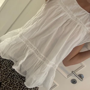 Vit blus med spetsdetaljer - Supersöt vit blus i luftig bomull med romantiska spetsdetaljer och volang nertill. Blusen är ärmlös och har en lös passform som ger en chill och somrig vibe. Perfekt till jeans eller kjol för en fräsch look.