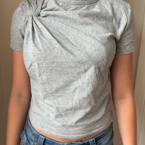 Grå T-shirt med en twist  - Säljer en ljusgrå croppad topp i bomull med korta ärmar och en snygg detalj längst upp! T-shirten, har aldrig använts. Säljer på grund av att den tyvärr inte passar mig. pris kan diskuteras!💖
