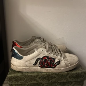 Gucci Ace sneakers med orm brodyr - Gucci Ace sneakers i vitt skinn med ikonisk röd och svart orm brodyr på sidan. Baksidan har metallicröd och metallicblå detalj med Gucci-logga. Klassisk rund tå och platt sula. En tvätt hade fått dom se nya ut!