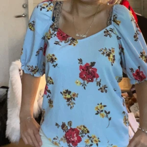 Blommig ljusblå blus med puffärm - Supersöt ljusblå blus i nyskick från Vila. Med röda och gula blommor, puffiga korta ärmar och djup urringning både fram och bak. Blusen har resår vid ärmslut och går ner lite i ryggen. Perfekt för dig som gillar romantisk och trendig stil.