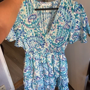 Blåvit kortklänning från Zara XS - Supersöt kortklänning från Zara i blått, vitt och lila med paisleyinspirerat mönster. Klänningen har v-ringning, volangdetaljer och korta, luftiga ärmar. Resår i midjan för en snygg passform. Perfekt för sommarens alla tillfällen!