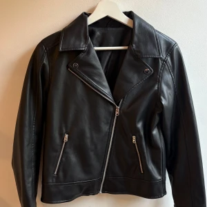 Svart skinnjacka med dragkedjor - Snygg svart skinnjacka med klassisk bikerstil, silvriga dragkedjor och bred krage. Jackan har två fickor framtill med dragkedja och en snedställd dragkedja för stängning. Perfekt för dig som vill ha en edgy och stilren look.