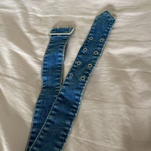 Brett jeansbälte med silverfärgat spänne - Säljer ett brett jeansbälte i klassisk blå denim med dubbla rader metallöljetter och ett stilrent, fyrkantigt silverfärgat spänne. Passar grymt till både jeans och kjol.