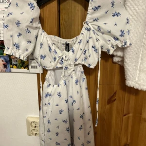 Vit blommig kortklänning H&M Divided - Supersöt vit kortklänning från H&M Divided med blått blommönster, puffärmar och smockad rygg. Klänningen har knytning framtill, cut-out vid midjan och voluminösa ärmar som ger en romantisk vibe. Perfekt för sommarens alla tillfällen!