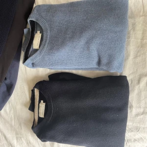 Blå och svart stickad tröja  - Två stilrena stickade tröjor, en i ljusblått och en i svart. Båda har rund halsringning och är ribbstickade vid kragen. Perfekta för lager-på-lager och passar till jeans eller kjol.