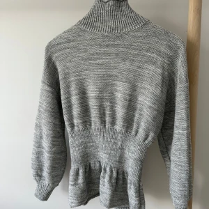 Grå stickad polotröja från SHEIN - Säljer en gråmelerad stickad polotröja från SHEIN. Tröjan har hög polokrage, markerad midja med ribbad struktur och volangliknande detalj nedtill. Långärmad och i mjukt material som passar perfekt när det är kyligt ute.