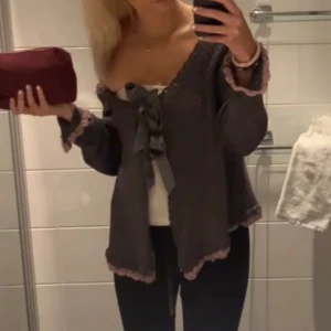 Grå stickad kofta med rosetter - Supersöt grå stickad kofta med ljusrosa virkade kanter och två satinrosetter framtill. Koftan har lång ärm, v-ringning och en loose passform. Perfekt för lager-på-lager och ger en romantisk vibe till outfiten.