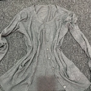 Grå kofta med knappar - En enkel och stilren grå kofta med långa ärmar och knappar hela vägen framtill. Koftan har en v-ringad halsringning och är tillverkad i ett mjukt, tunt stickat material som känns skönt mot huden. Perfekt att slänga över en t-shirt eller linne.