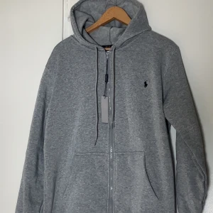 Grå hoodie från Polo Ralph Lauren - Snygg grå hoodie från Polo Ralph Lauren med dragkedja framtill och klassisk broderad logga på bröstet. Tröjan har huva med snörning, två fickor och är tillverkad i mjuk bomullsmix. Perfekt för en chill och stilren look.