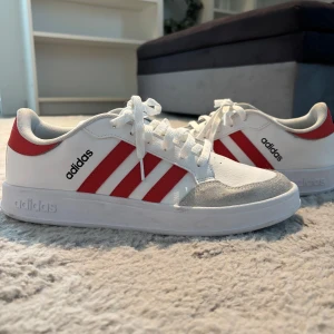 Adidas Breaknet Plus red/white - Fräscha vita Adidas sneakers med klassiska röda stripes på sidorna och grå mockadetaljer vid tån. Skorna har snörning och en platt sula med Adidas-logga undertill. Perfekt för dig som gillar en sportig och stilren look.