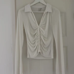 Vit omlottopp med rynk och knappar från Nelly - Säljer en vit långärmad topp från Gina Tricot med snygg rynk framtill, djup v-ringning och knappar längs hela framsidan. Tunt och mjukt material, perfekt till jeans eller kjol. Krage för extra stilpoäng!