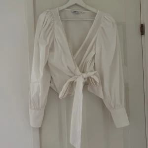 Vit omlottblus med knyt från Kappahl - Supersnygg vit omlottblus från Kappahl med puffiga långärmar och breda manschetter. Blusen har v-ringning och knyts i midjan för en trendig look. Perfekt att styla med jeans eller kjol för en chic vibe.