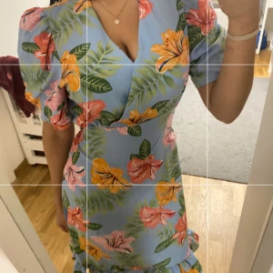 Blommig blå klänning Paris Et Moi - Superfin blå klänning från Paris Et Moi med färgglada blommor i rosa, orange och grönt. Klänningen har puffärmar, v-ringning och volang nertill. Perfekt för sommarens alla tillfällen och ger en riktigt fräsch vibe.