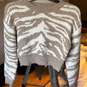 Beige stickad tröja - Säljer en beige och vit stickad tröja från Shein med zebramönster. Tröjan är croppad med ribbade muddar och rund hals. Perfekt för dig som vill ha en trendig och mysig look till hösten eller vintern. Storlek M🌸