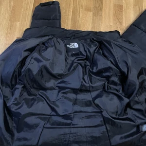 Svart pufferjacka från The North Face - Säljer en svart pufferjacka från The North Face i storlek L. Jackan har en klassisk pufferdesign med diskret logga på bröstet, dragkedjor på fickorna och hög krage. Perfekt för kalla dagar när du vill hålla stilen och värmen.