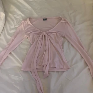 Ljusrosa omlottopp från Gina Tricot - Supersöt ljusrosa omlottopp från Gina Tricot med långa ärmar och knyt framtill. Toppens material är mjukt och stretchigt, perfekt för en trendig och feminin look. Djup v-ringning och smickrande passform som framhäver midjan.