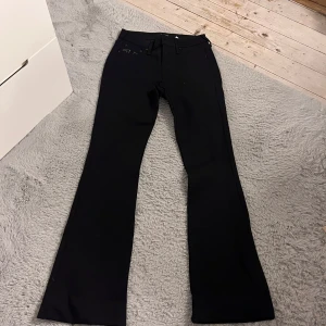 Svarta Mid Rise bootcut True Religion - Svarta kostym byxor från True Religion med bootcut passform och klassiska kontrastsömmar på bakfickorna. Snyggt broderad logga på fickorna och patch med varumärket bak i midjan. Perfekta för en trendig look.