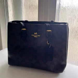 Svart handväska med mönster - Stilren svart handväska med mörkt mönster och guldfärgade detaljer. Väskan har två handtag, justerbara remmar och en tag med logga. Tillverkad i skinnimitation och har en klassisk, fyrkantig form. Perfekt för dig som gillar snygga accessoarer. AA copy