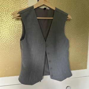Grå klassisk kostymväst - Stilren grå väst med knappar framtill och klassisk v-ringning. Perfekt för att lyfta en skjorta eller t-shirt. Västen har en figurnära passform och är tillverkad i ett slätt tyg som ger en dressad look.