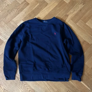 Mörkblå sweatshirt från Polo Ralph Lauren - Klassisk mörkblå sweatshirt från Polo Ralph Lauren med rund halsringning och röd broderad logga på bröstet. Tröjan har långa ärmar och ribbade muddar vid ärmslut och nederkant. Perfekt för en chill och stilren look.