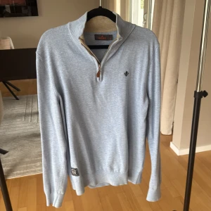 Ljusblå half zip tröja från Morris - Snygg ljusblå tröja från Morris med half zip och ribbad krage. Tröjan har lång ärm, diskret broderad logga på bröstet och är tillverkad i mjuk bomull. Perfekt för dig som gillar stilrena och klassiska plagg med en modern touch.