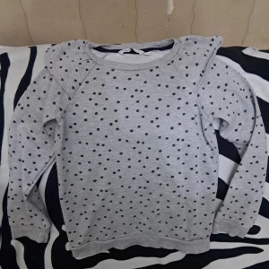 Grå prickig sweatshirt med puffärm (barn) - Grå sweatshirt med svarta prickar och coola puffärmar. Tröjan har rund halsringning och långa ärmar, perfekt för dig som gillar en lekfull men stilren look. Mjuk och skön, passar till jeans eller kjol för en chill vibe. Den är i väldigt fint skick :) (barn tröja) - storlek 134/140