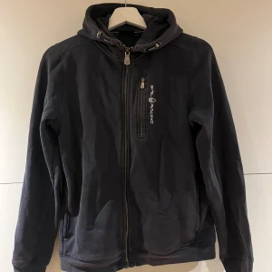 Blå hoodiejacka från Sail Racing - Blå hoodiejacka från Sail Racing med dragkedja framtill och bröstficka med logga. Jackan har huva med snörning och långa ärmar. Perfekt för dig som gillar en sportig och clean stil.