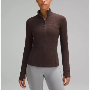 Lululemon define jacket - Slimmad brun jacka från Lululemon med dragkedja framtill och höga muddar med tumhål. Jackan har en sportig look, figurnära passform och hög krage. Perfekt för dig som gillar en clean och stilren stil.