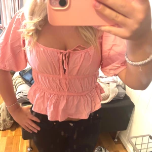 Rosa volangblus med puffärm och knyt - Supersöt rosa blus med volangkant och puffiga korta ärmar. Toppen har smockad midja och knyt framtill vid bysten för extra detalj. Perfekt för dig som gillar en feminin och trendig look. Croppad modell som passar till både jeans och kjol.