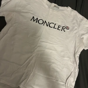 Vit Moncler t-shirt med logga - Snygg vit t-shirt från Moncler i bomull med stor svart Moncler-logga tryckt på bröstet och klassisk rund hals. Perfekt för dig som gillar stilrena och exklusiva plagg med tydlig branding.