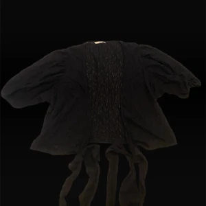 Svart spetsig knytblus från H&M - Svart blus från H&M med puffiga ärmar och vacker spets framtill. Modellen har knytband i midjan och är tillverkad i ett lätt, transparent tyg som ger en cool och luftig känsla. Perfekt för dig som gillar detaljer och en unik look.