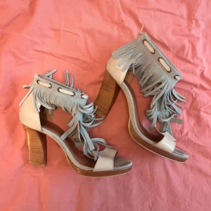 Ljusblå mockaklackar med fransar - Snygga sandaler i ljusblå mocka med höga klackar och coola fransdetaljer runt ankeln. Skorna har öppen tå, dragkedja bak och en stadig träfärgad klack. Perfekta för dig som vill sticka ut med en boho-vibe och älskar detaljer.