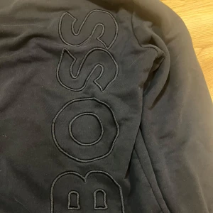 Svart sweatshirt från BOSS - Svart sweatshirt från BOSS med stort broderat logotyptryck längs framsidan. Tröjan har rund halsringning, långa ärmar och ribbade muddar vid ärmslut och nederkant. Perfekt för en stilren och avslappnad look.