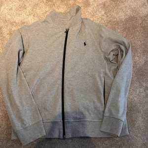 Grå zipjacka från Polo Ralph Lauren - Snygg grå zipjacka från Polo Ralph Lauren med svart broderad logga på bröstet. Jackan har hög krage, hel dragkedja och är tillverkad i mjuk bomull. Perfekt för en chill och stilren look.