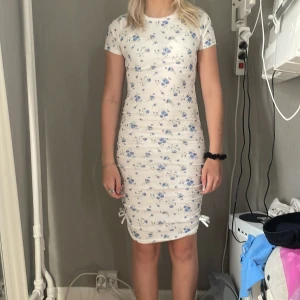 Vit blommig kortklänning från Shein - Supersöt vit kortklänning från Shein med blått blommönster. Klänningen är ribbad, har korta ärmar och snörning i sidorna för en extra snygg detalj. Perfekt för sommarens alla tillfällen och sitter tight och bekvämt.