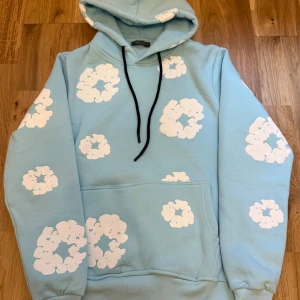 Ljusblå hoodie från Denim Tears - Säljer en ljusblå hoodie från Denim Tears med vita blommiga tryck över hela tröjan. Tröjan har huva med svarta snören och en stor magficka. Perfekt för dig som gillar streetwear och vill sticka ut med en unik design.