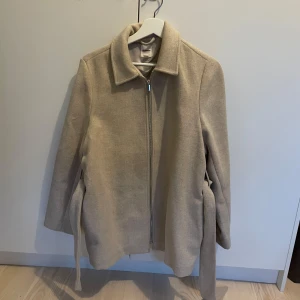 Beige kappa med bälte från Dobber - Stilren beige kappa från Dobber med dragkedja framtill och bälte i midjan. Aldrig använd 