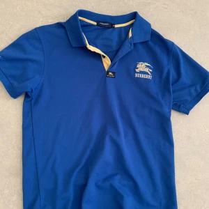 Blå pikétröja från Burberry - Snygg blå pikétröja från Burberry med gulddetaljer på kragen och broderad logga både fram och bak. Klassisk pikékrage med knappslå och korta ärmar. Perfekt för dig som vill ha en stilren och exklusiv look.