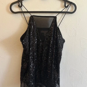 Svart paljettlinne med tunna band - Glittrig svart linnetopp med tunna axelband och v-ringning. Hela toppen är täckt av svarta paljetter som ger en riktigt snygg partykänsla. Sitter löst och har en lätt croppad look. Perfekt att styla med jeans eller kjol för en extra glammig outfit.