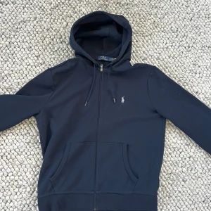 Mörkblå hoodie från Polo Ralph Lauren - Snygg mörkblå hoodie från Polo Ralph Lauren med dragkedja framtill och klassisk logga broderad på bröstet. Tröjan har huva med snörning och två stora fickor framtill. Perfekt för en chill och stilren look.