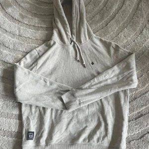 Ljusgrå hoodie från Morris - Snygg ljusgrå hoodie från Morris med broderad logga på bröstet och patch nertill. Klassisk huva med snörning och långa ärmar. Mjuk bomullsblandning som är perfekt för chill dagar. Passar dig som gillar stilren och enkel design.