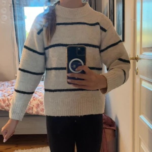 Randig stickad tröja från Gina Tricot - Mysig stickad tröja från Gina Tricot i ljusgrått med svarta horisontella ränder. Tröjan har en hög, ribbad halsringning och långa ärmar. Perfekt för kyliga dagar och enkel att matcha med jeans eller leggings.