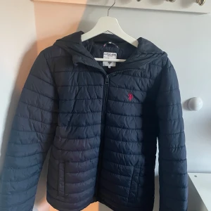   Polo jacka helt ny - Mörkblå pufferjacka från U.S. Polo Assn med huva och dragkedja framtill. Jackan har quiltad design och röd logga broderad på bröstet. Perfekt för kyliga dagar och har en clean, sportig look.